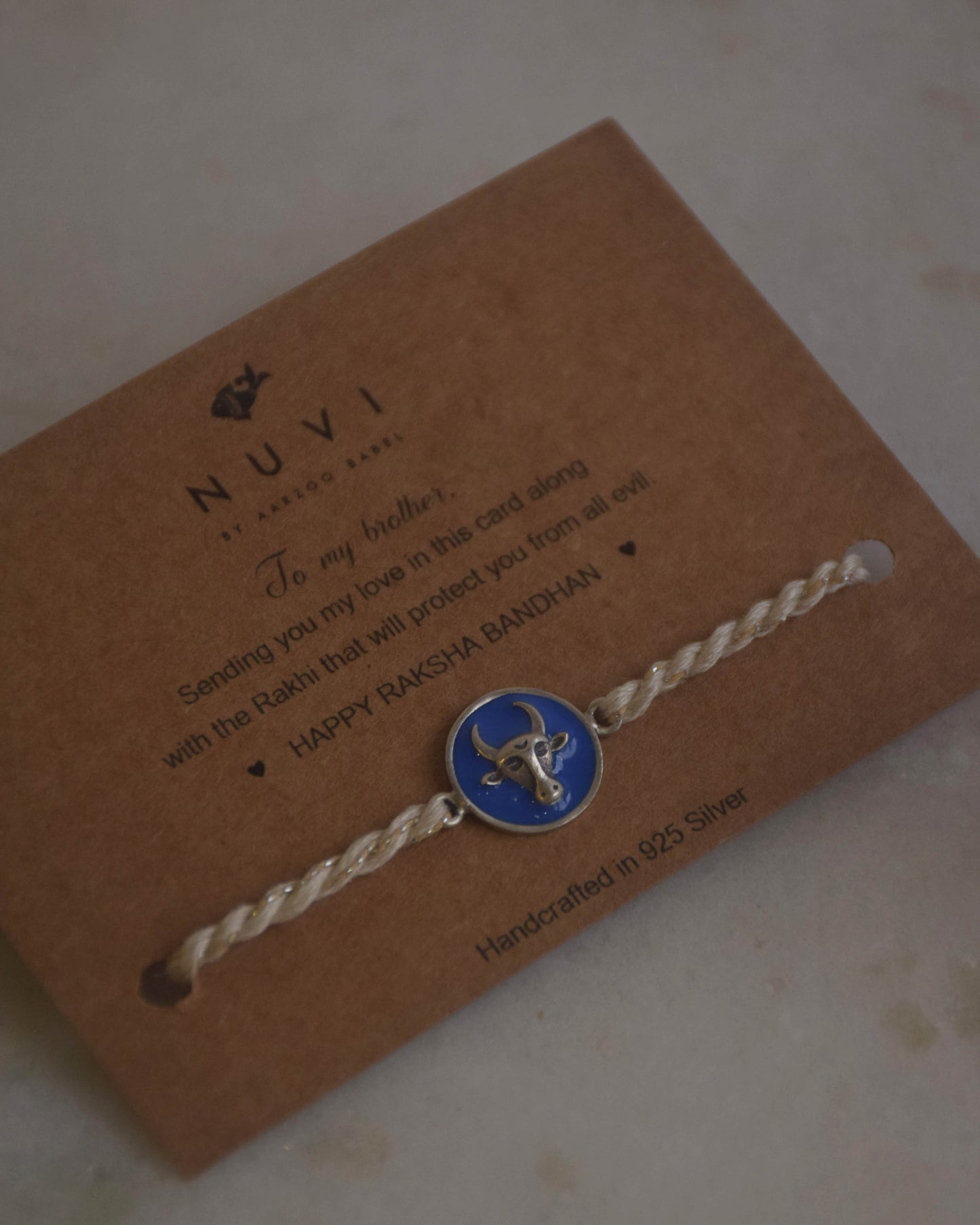 925 Silver Rakhi with blue Nandi motif on a card with 'NUVI' branding, placed on a textured surface.