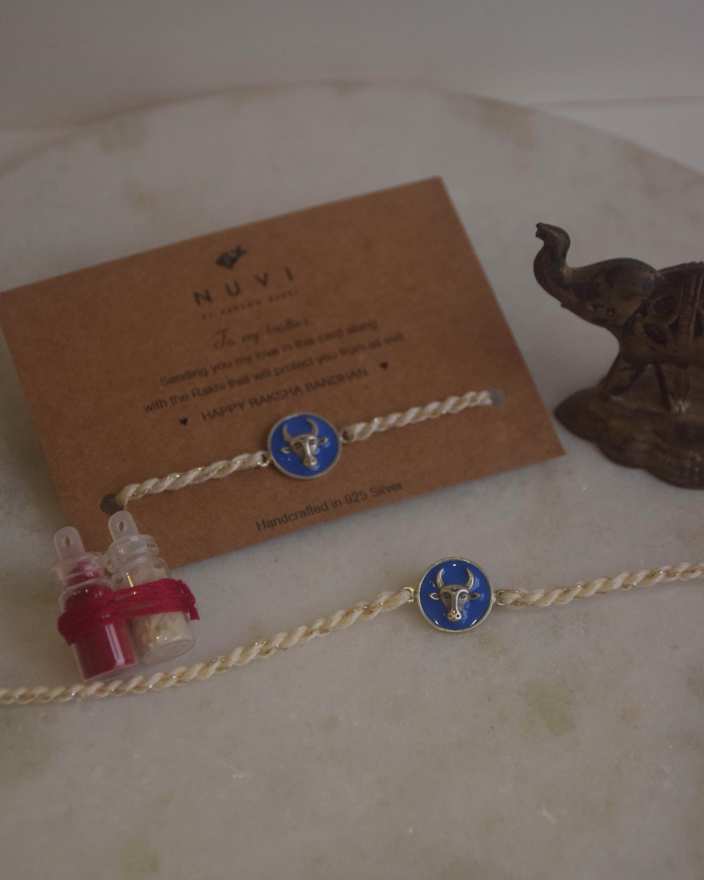 925 Silver Rakhi with blue enamel and Nandi motif on a card with 'NUVI' branding, placed on a textured surface.