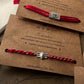 Two sterling silver Rakhi with red cotton thread on brown cards with 'NUVI' branding and text.