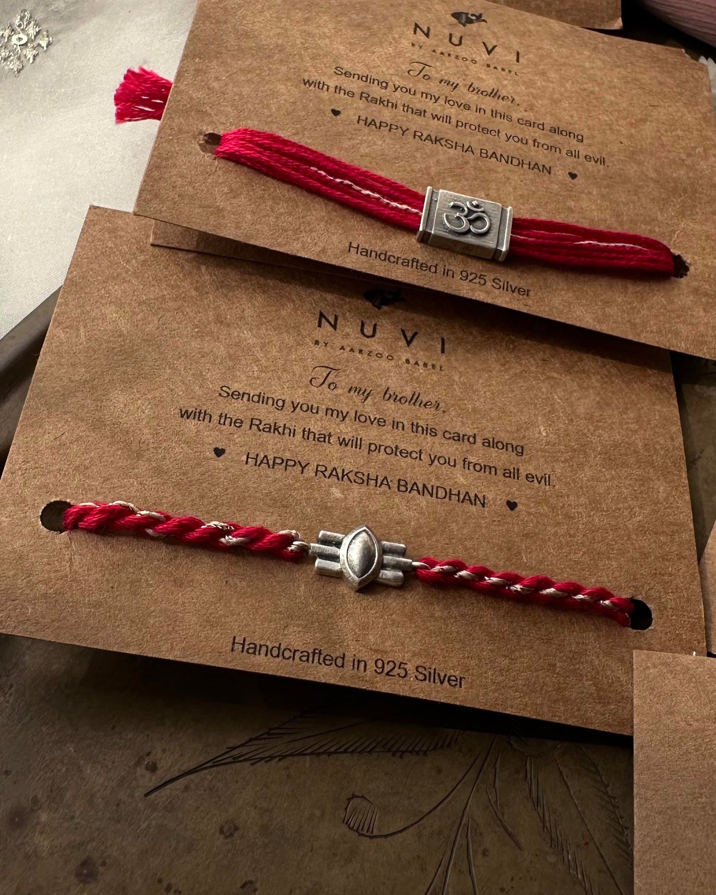 Two sterling silver Rakhi with red cotton thread on brown cards with 'NUVI' branding and text.