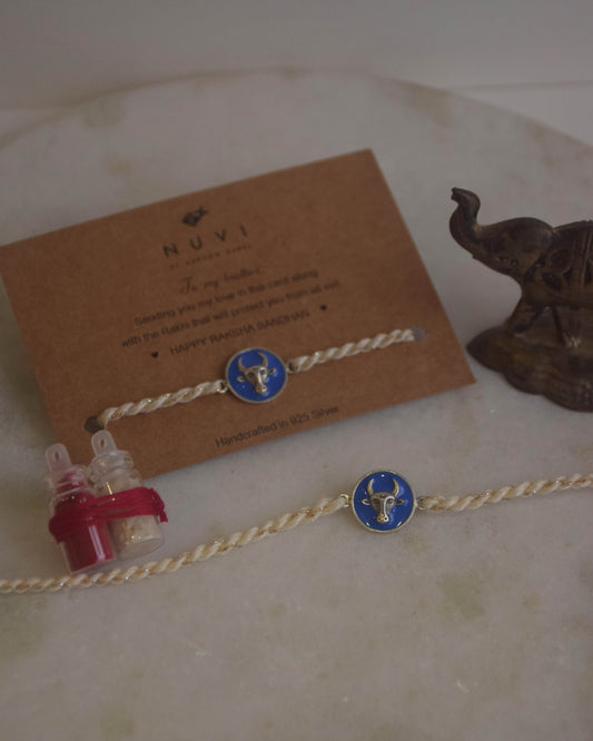 925 Silver Rakhi with blue enamel and Nandi motif on a card with 'NUVI' branding, placed on a textured surface.
